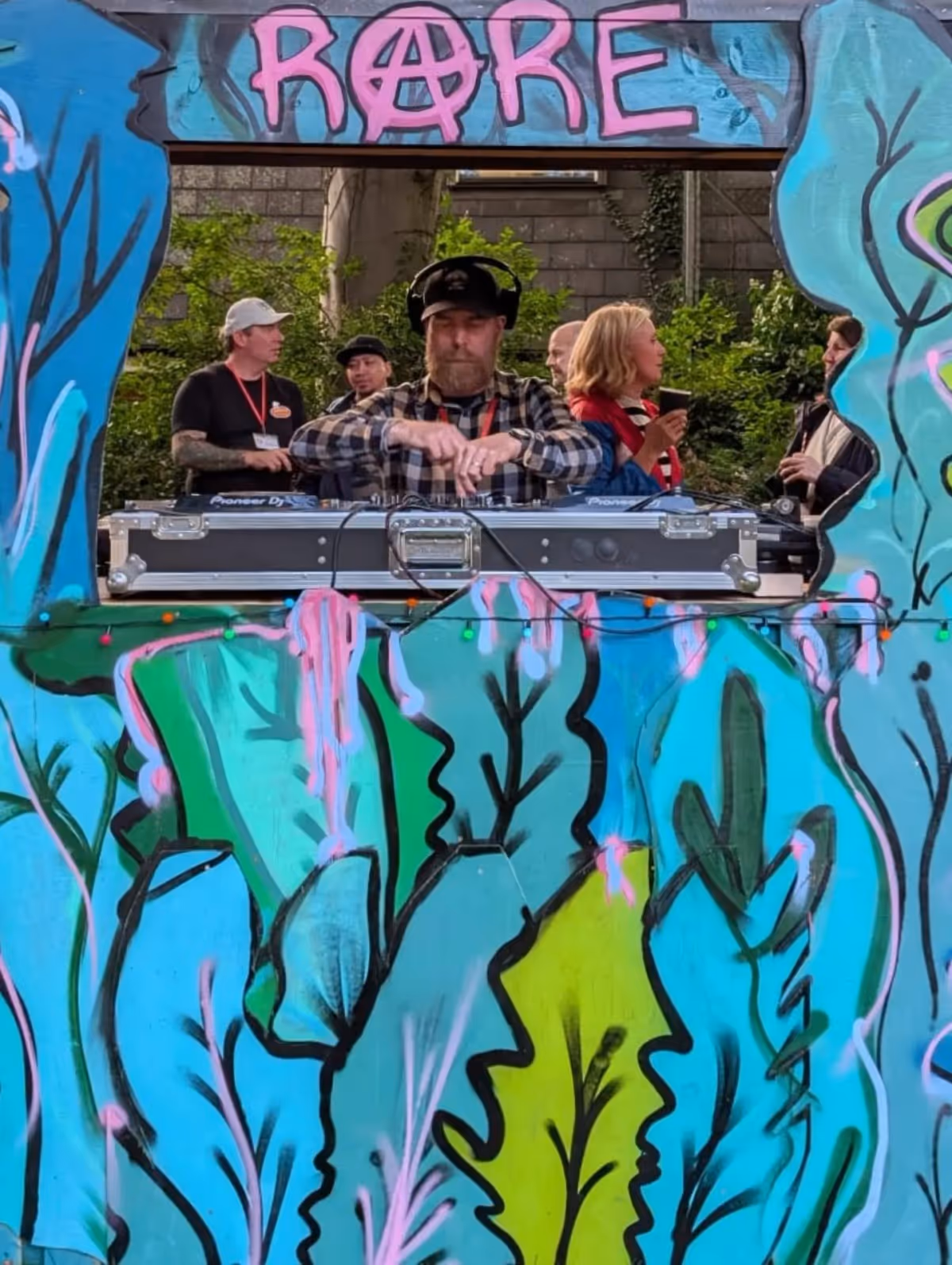 Smiley Jack wearing headphones behind the decks in a colourful outdoor RARE DJ booth.